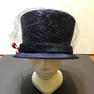 May include: A navy blue hat with a black net veil and a red ribbon. The hat is sitting on a white mannequin head.