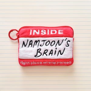 May include: A red and white rectangular pouch with the words "INSIDE NAMJOON'S BRAIN" embroidered in white and black. The pouch has a red zipper and a red ring attached to the side. The text "right place wrong person" is at the bottom.