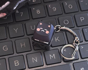 May include: A black mechanical keyboard keycap keychain featuring a stylized cat face with pink cheeks, a small red horizontal line, and a closed-eye expression. The keycap is attached to a silver-toned metal split ring by a short coiled wire.