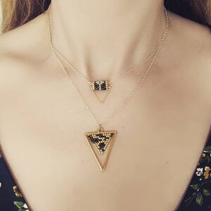 Puede incluir: Un collar en capas con dos colgantes. El colgante superior es un pequeño triángulo dorado con un centro de cuentas. El colgante inferior es un triángulo dorado más grande con un centro de cuentas. El collar lo lleva una persona.
