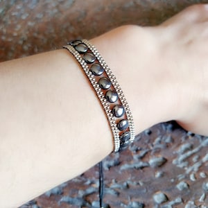 May include: A dark grey bracelet featuring a series of dark grey, circular stones. The bracelet is made of a silver-coloured metal and is worn on a wrist. The bracelet is secured with a black string.