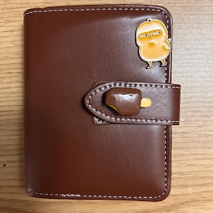 Puede incluir: Una cartera de piel sintética marrón con cierre de broche decorado con un dije de helado cubierto de chocolate. La parte frontal de la cartera presenta un pequeño pin esmaltado de un pollito amarillo con expresión gruñona.
