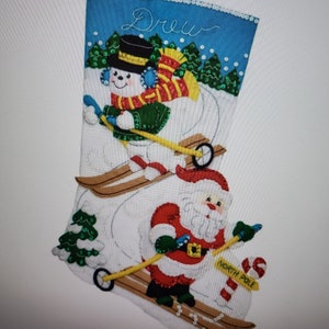 May include: A white felt Christmas stocking with a blue background and the name "Drew" embroidered on it. The stocking features a snowman and Santa Claus skiing down a snowy hill with a green Christmas tree in the background. The stocking has a red loop at the top for hanging.