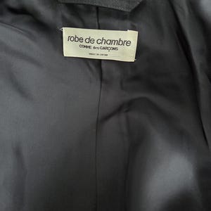 Puede incluir: Primer plano del interior de una prenda gris oscuro. Una etiqueta rectangular est&aacute; cosida en el forro, mostrando el texto "robe de chambre COMME des GAR&Ccedil;ONS MADE IN JAPAN" en una fuente serif. La tela parece lisa y mate.