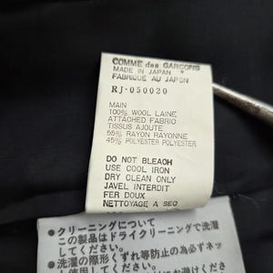 Puede incluir: Primer plano de dos etiquetas de tela blancas con texto negro. La etiqueta superior dice "COMME des GARCONS MADE IN JAPAN FABRIQUE AU JAPON." La etiqueta inferior tiene texto japon&eacute;s e instrucciones de cuidado.