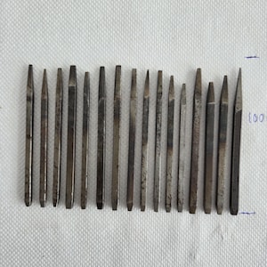 Puede incluir: Una colección de herramientas metálicas antiguas, probablemente punzones o cinceles, dispuestas en fila. Las herramientas tienen forma rectangular o cónica con un acabado metálico opaco. Están colocadas sobre una superficie blanca texturizada.