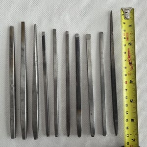 Puede incluir: Un conjunto de herramientas metálicas, incluyendo cinceles y punzones, dispuestas sobre una superficie blanca. Una cinta métrica amarilla se encuentra junto a las herramientas, mostrando medidas en centímetros. Las herramientas varían en tamaño y forma.