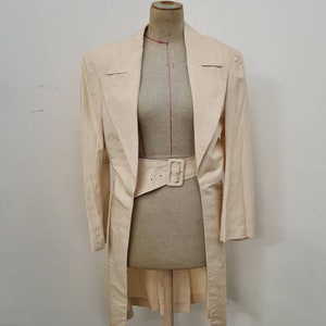 May include: A long, off-white linen blazer with a wide, brown belt. The blazer has a shawl collar and a single-breasted closure.