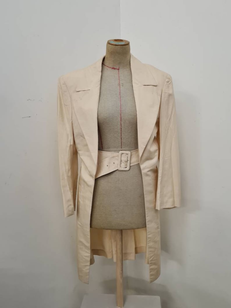 May include: A long, off-white linen blazer with a wide, brown belt. The blazer has a shawl collar and a single-breasted closure.