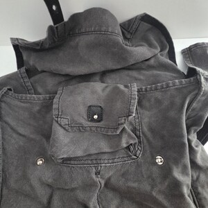 May include: Close-up of a charcoal-colored jacket with a hood and multiple pockets. The jacket features a small square pocket with a button closure and two silver-colored buttons. A black strap is visible at the top.