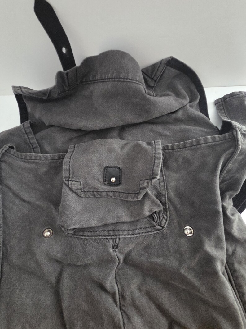 May include: Close-up of a charcoal-colored jacket with a hood and multiple pockets. The jacket features a small square pocket with a button closure and two silver-colored buttons. A black strap is visible at the top.