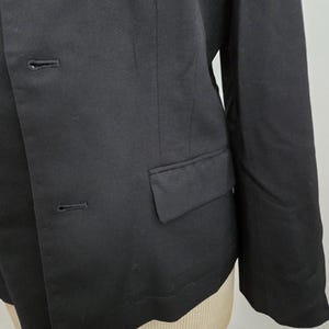 Puede incluir: Primer plano de una blazer negra con dos botones y un bolsillo. La blazer est&aacute; hecha de una tela lisa y tiene un corte entallado. La manga es visible y la prenda se muestra en un maniqu&iacute;.