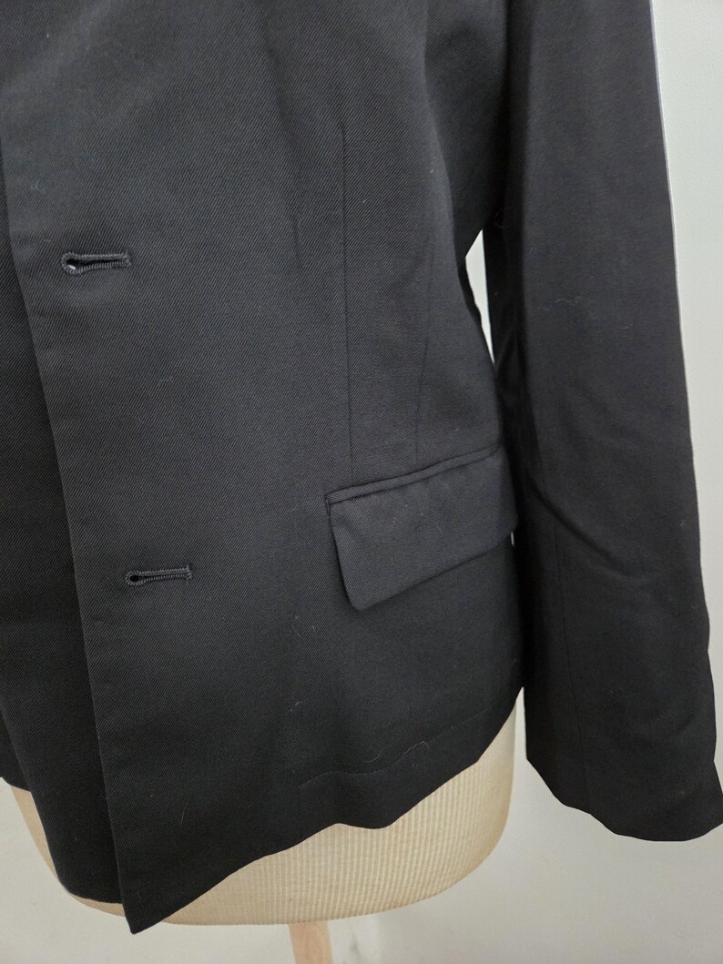 Puede incluir: Primer plano de una blazer negra con dos botones y un bolsillo. La blazer est&aacute; hecha de una tela lisa y tiene un corte entallado. La manga es visible y la prenda se muestra en un maniqu&iacute;.
