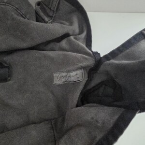 May include: A dark gray hooded sweatshirt with a faded appearance. The hood is up, and the fabric is textured. A small rectangular label with cursive text is visible on the chest.