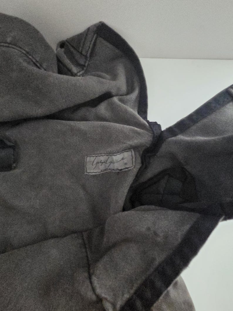 May include: A dark gray hooded sweatshirt with a faded appearance. The hood is up, and the fabric is textured. A small rectangular label with cursive text is visible on the chest.