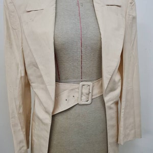 May include: A long, off-white blazer with a wide, brown belt. The blazer has a shawl collar and long sleeves.