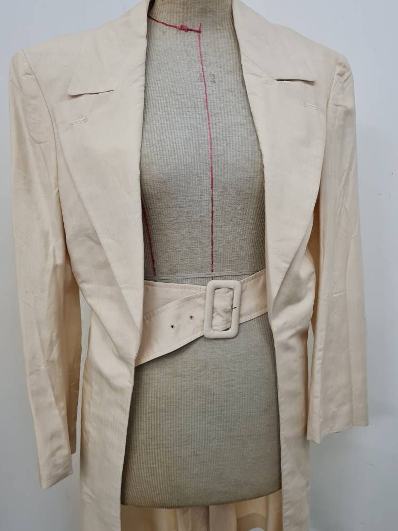 May include: A long, off-white blazer with a wide, brown belt. The blazer has a shawl collar and long sleeves.