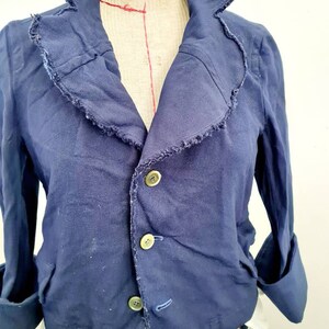 May include: A navy blue cropped jacket with a ruffled collar and three button closures. The jacket has long sleeves with rolled cuffs.