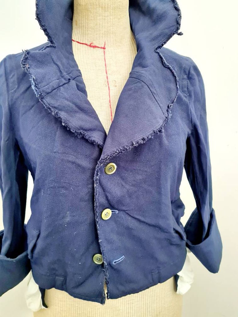 May include: A navy blue cropped jacket with a ruffled collar and three button closures. The jacket has long sleeves with rolled cuffs.