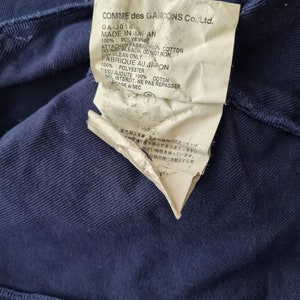 May include: A white tag with black text attached to a navy blue garment. The tag reads "COMME des GARCONS Co., Ltd. GA-J016 MADE IN JAPAN 100% POLYES ATTACHED FABRIC 100% COTTON DO NOT BLEACH, DO NOT IRON. DRY CLEAN ONLY. FABRIQUE AU JAPON 100% POLYESTER TISSU AJOUTE 100% COTON NEI INTERDIT, NE PAS REPASSER. AGE A SEC."