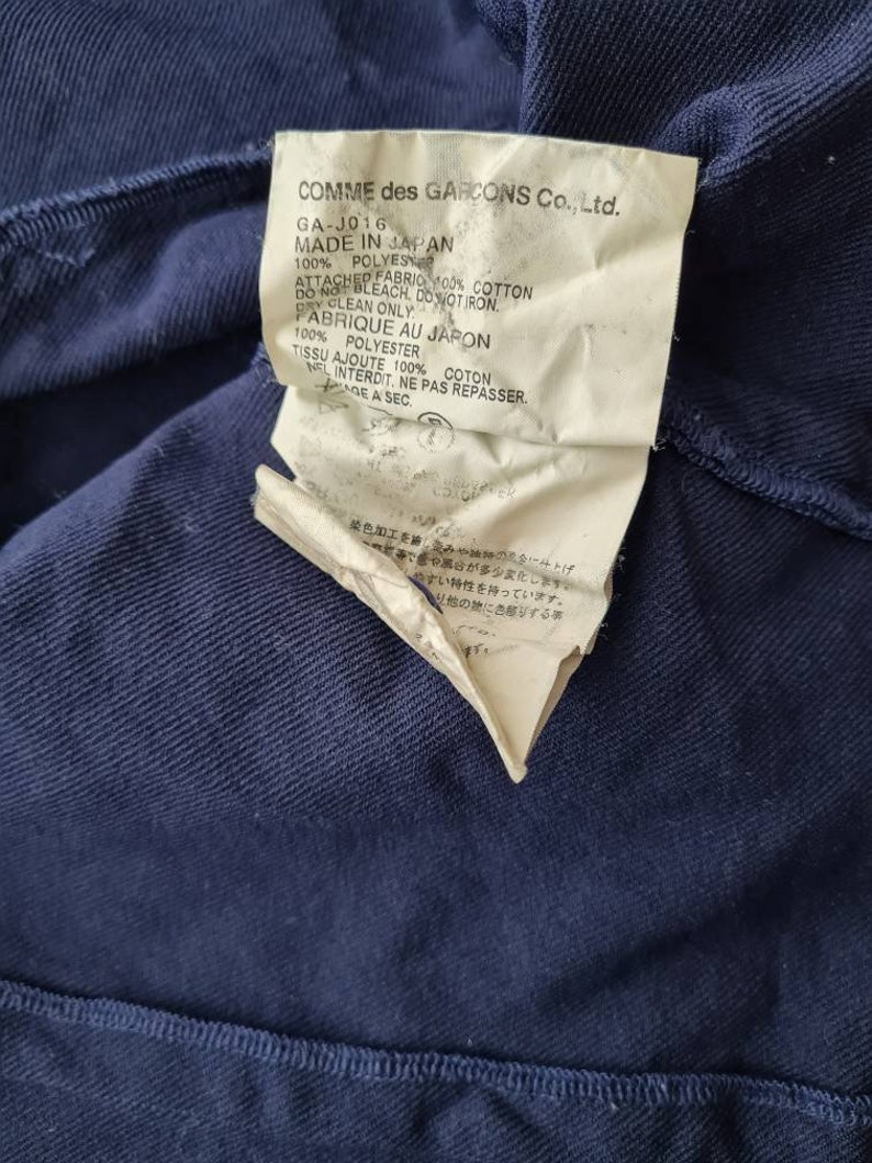 May include: A white tag with black text attached to a navy blue garment. The tag reads "COMME des GARCONS Co., Ltd. GA-J016 MADE IN JAPAN 100% POLYES ATTACHED FABRIC 100% COTTON DO NOT BLEACH, DO NOT IRON. DRY CLEAN ONLY. FABRIQUE AU JAPON 100% POLYESTER TISSU AJOUTE 100% COTON NEI INTERDIT, NE PAS REPASSER. AGE A SEC."
