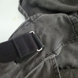May include: Close-up of a dark gray canvas bag with a black strap and silver-colored metal hardware. The bag's fabric appears worn, with visible stitching and a textured surface. The strap is made of a woven material.