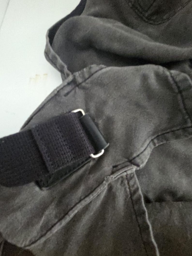 May include: Close-up of a dark gray canvas bag with a black strap and silver-colored metal hardware. The bag's fabric appears worn, with visible stitching and a textured surface. The strap is made of a woven material.