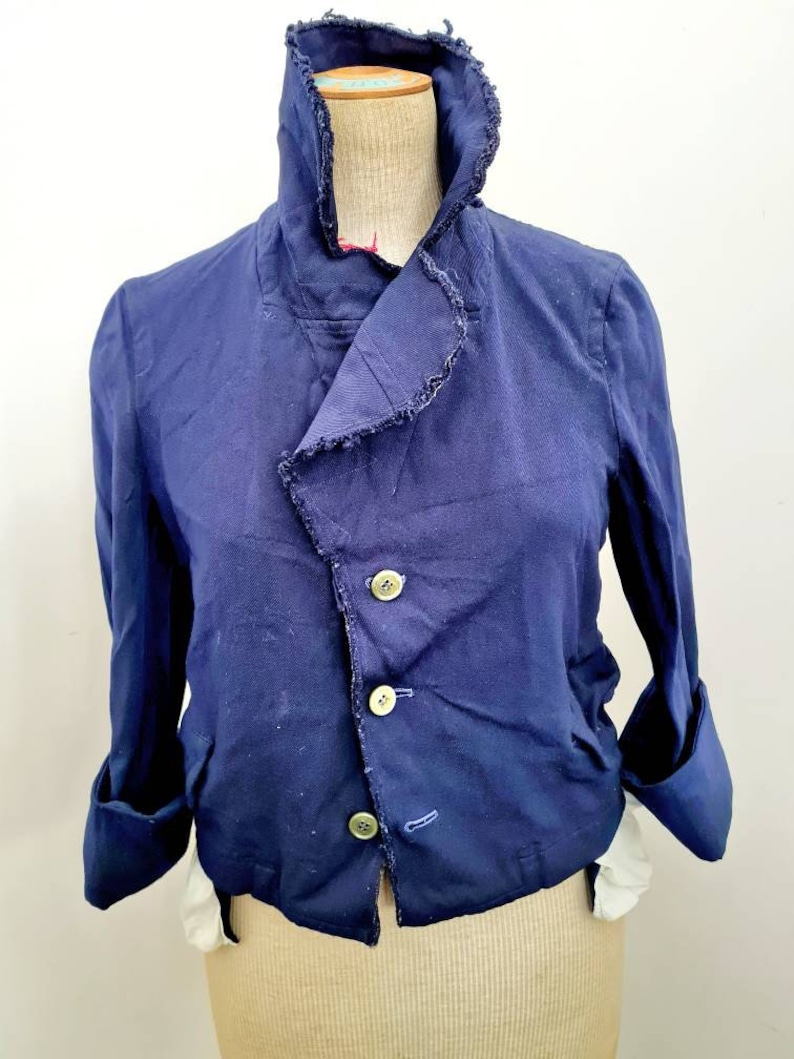 May include: A navy blue cropped jacket with a frayed collar and three gold buttons. The jacket has long sleeves with rolled up cuffs.
