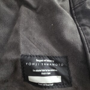 May include: Close-up of a dark gray garment with a black label. The label reads "Designed and tailored by YOHJI YAMAMOTO" with additional text. The garment appears to be made of a soft, textured fabric.