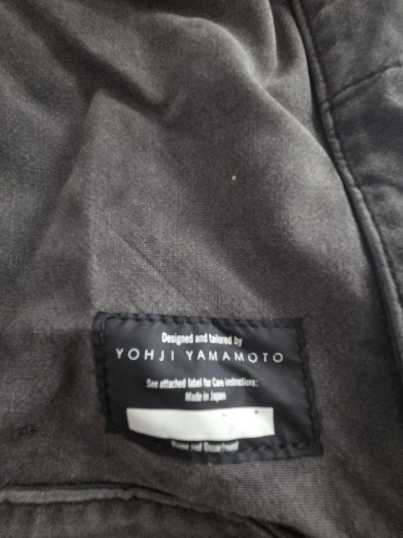 May include: Close-up of a dark gray garment with a black label. The label reads "Designed and tailored by YOHJI YAMAMOTO" with additional text. The garment appears to be made of a soft, textured fabric.