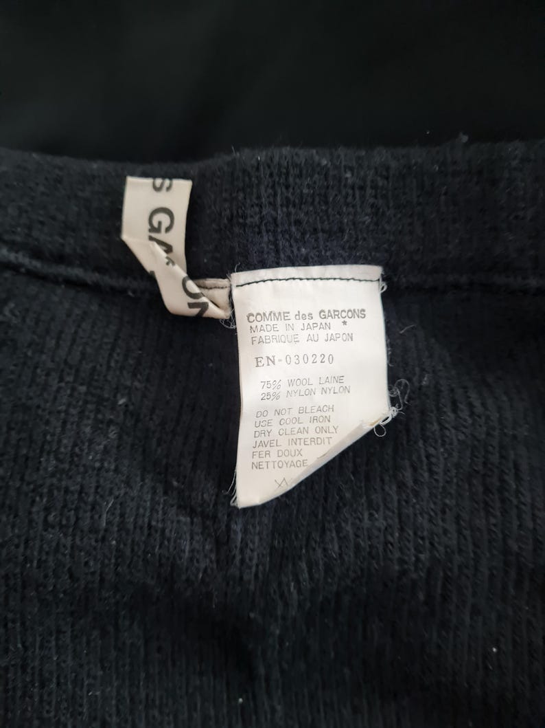 Pode incluir: Um su&eacute;ter de malha preto com uma etiqueta branca que diz "COMME des GARCONS MADE IN JAPAN FABRIQUE AU JAPON EN-030220 75% WOOL LAINE 25% NYLON NYLON DO NOT BLEACH USE COOL IRON DRY CLEAN ONLY JAVEL INTERDIT FER DOUX NETTOYAGE"