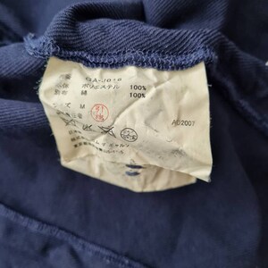 May include: A white tag with black text attached to a dark blue garment. The tag reads "GA-J016", "Polyester 100%", "Lining: Cotton 100%", "Size M", "AD2007".