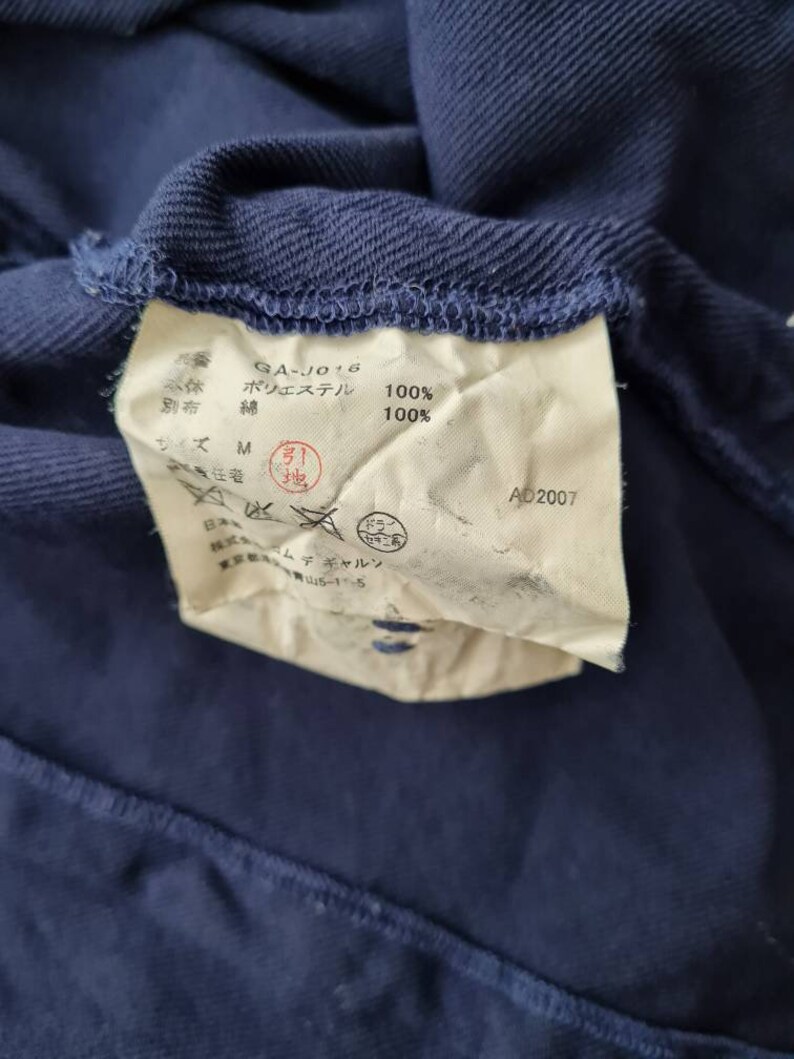 May include: A white tag with black text attached to a dark blue garment. The tag reads "GA-J016", "Polyester 100%", "Lining: Cotton 100%", "Size M", "AD2007".
