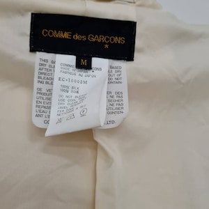 May include: A black and white care label for a Comme des Gar&ccedil;ons garment. The label is labelled with a size M and the text "Made in Japan. Fabrique au Japon." The label also includes the text "100% Silk. 100% Soie" and care instructions.