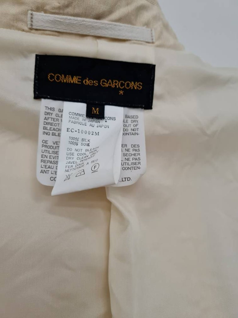 May include: A black and white care label for a Comme des Gar&ccedil;ons garment. The label is labelled with a size M and the text "Made in Japan. Fabrique au Japon." The label also includes the text "100% Silk. 100% Soie" and care instructions.