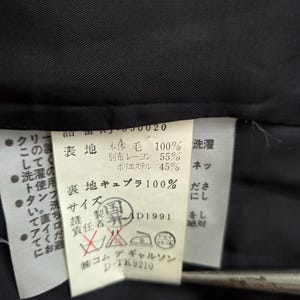 Puede incluir: Primer plano de una etiqueta blanca con texto y s&iacute;mbolos japoneses, adherida a una tela negra. La etiqueta incluye instrucciones de lavado y detalles de la composici&oacute;n de la tela.