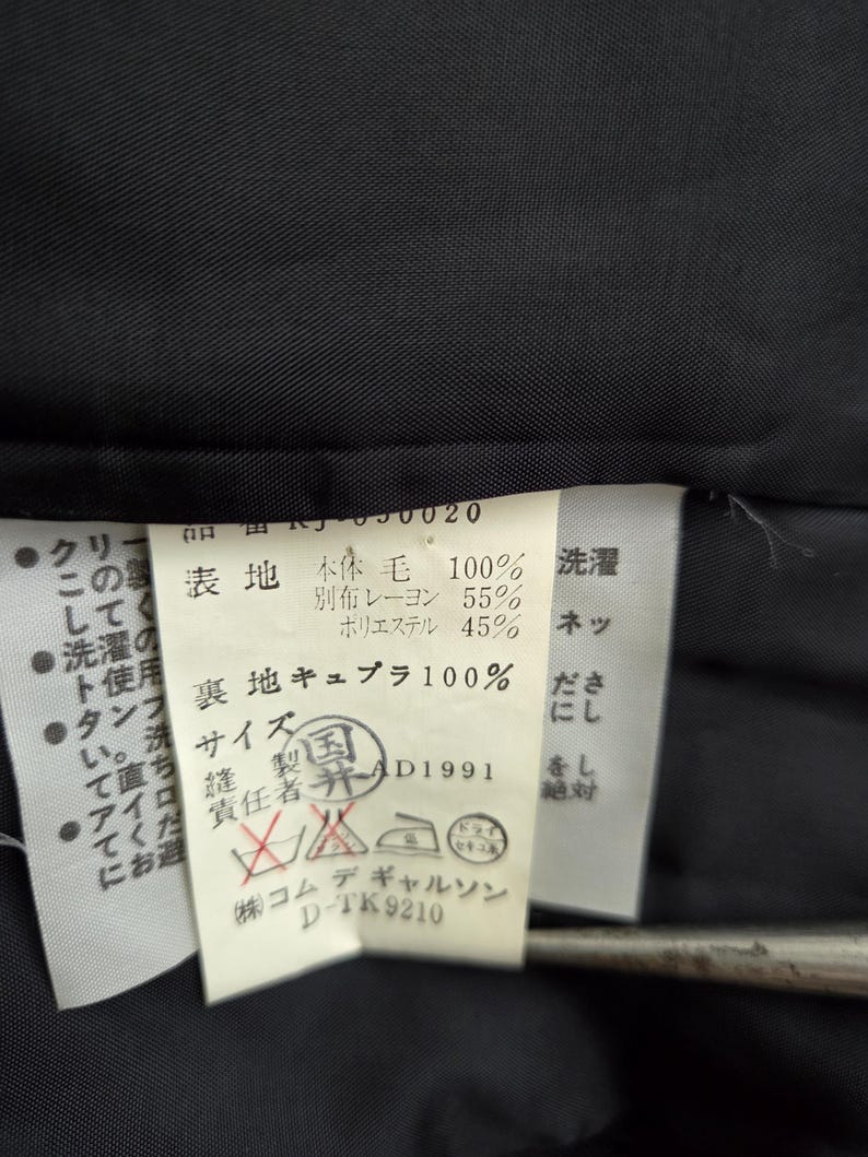 Puede incluir: Primer plano de una etiqueta blanca con texto y s&iacute;mbolos japoneses, adherida a una tela negra. La etiqueta incluye instrucciones de lavado y detalles de la composici&oacute;n de la tela.