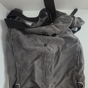 May include: A gray canvas messenger bag with a black adjustable shoulder strap. The bag has a rectangular shape with rounded corners and metal D-rings. The fabric appears worn, giving it a vintage look.