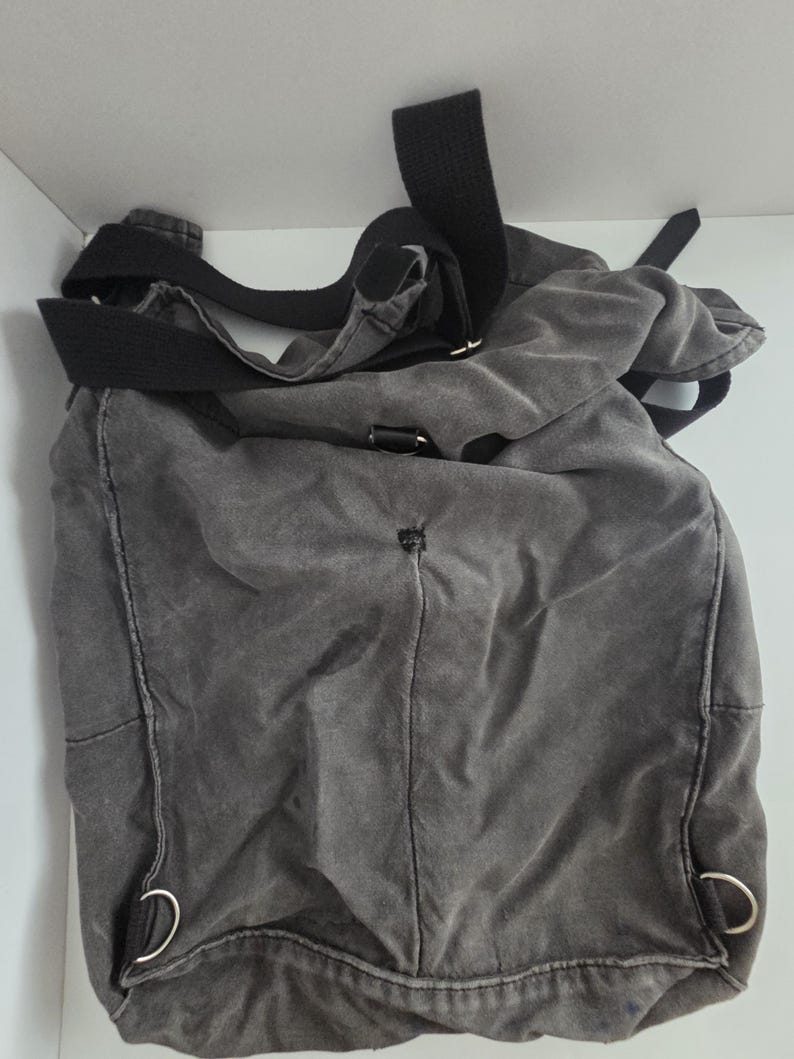 May include: A gray canvas messenger bag with a black adjustable shoulder strap. The bag has a rectangular shape with rounded corners and metal D-rings. The fabric appears worn, giving it a vintage look.