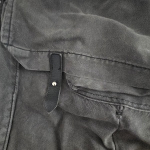 May include: Close-up of a gray canvas bag with a black leather strap and silver button closure. The bag has a pocket and visible stitching details. The fabric appears worn and textured.