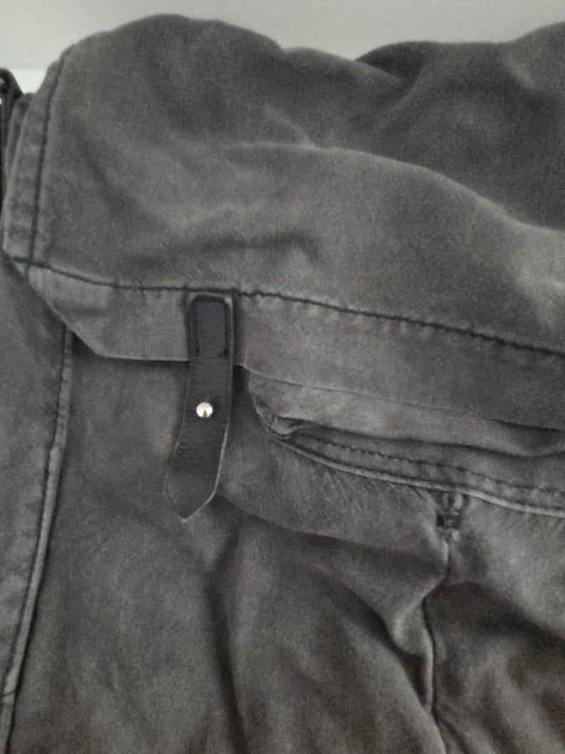 May include: Close-up of a gray canvas bag with a black leather strap and silver button closure. The bag has a pocket and visible stitching details. The fabric appears worn and textured.