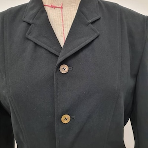 Puede incluir: Una chaqueta negra con cuello de solapa y cuatro botones dorados. La chaqueta está hecha de una tela lisa y texturizada.