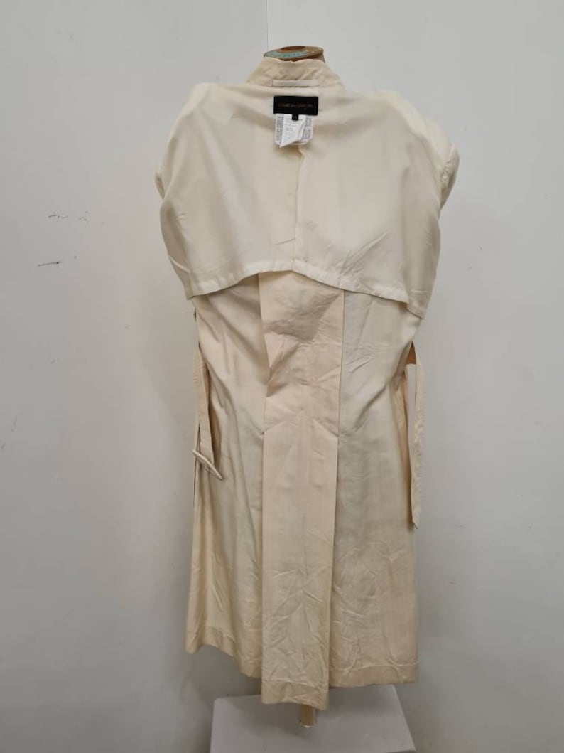 May include: A cream-coloured trench coat with a long, flowing silhouette. The coat has a belt and a collar. The label on the back of the coat reads "Gianfranco Ferre".