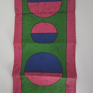 May include: A rectangular textile with a repeating pattern of circles and rectangles. The design features pink, green, and blue colours. The textile has a textured appearance and a small text in the bottom left corner.