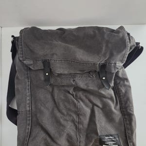 May include: A gray canvas messenger bag with a flap closure and black leather straps. The bag has a rectangular shape with a front pocket and a shoulder strap. The label reads "YOHJI YAMAMOTO".