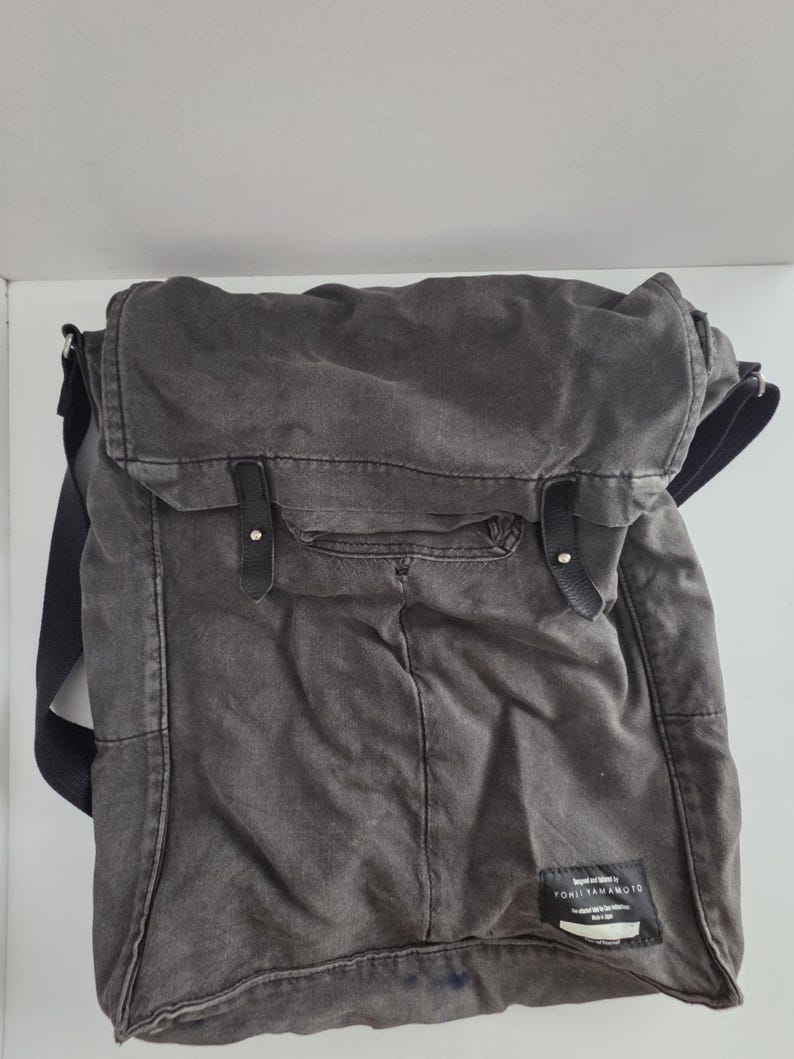 May include: A gray canvas messenger bag with a flap closure and black leather straps. The bag has a rectangular shape with a front pocket and a shoulder strap. The label reads "YOHJI YAMAMOTO".