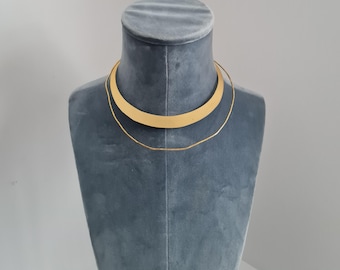 May include: A gold choker necklace with a wider, curved band and a thinner chain layered on top.