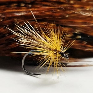 May include: Close-up of a fishing fly with a curved hook. The fly features a yellow and brown feathery design, with a dark head and iridescent accents. The background is a blurred arrangement of brown feathers.
