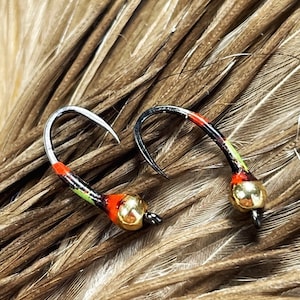 May include: Two fishing flies with curved silver hooks, black bodies, and colorful accents of red, green, and yellow. Each fly has a gold-colored bead at the base. The flies rest on a bed of brown feathers.