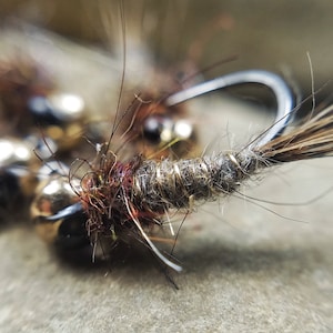 May include: Close-up of a fishing fly with a curved hook. The fly features a body wrapped in gray thread, with brown and tan feather accents. Small, shiny beads are visible near the hook.